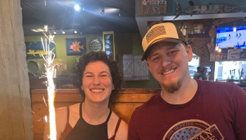 A smiling couple posing at a restaurant booth with a bright sparkler burning beside them.