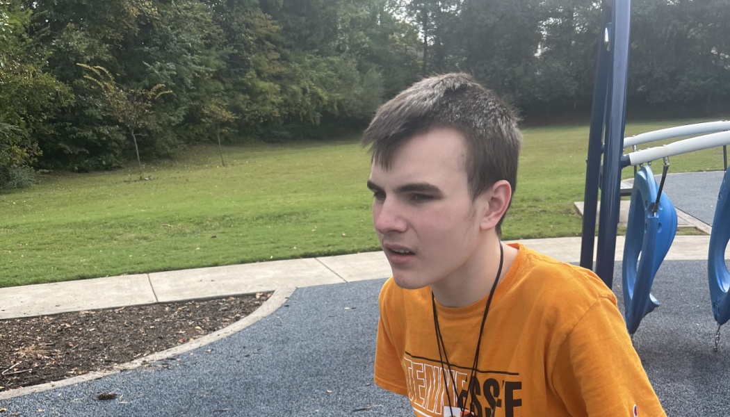 A young man in an orange t-shirt looking off-camera while sitting at an outdoor park playground.