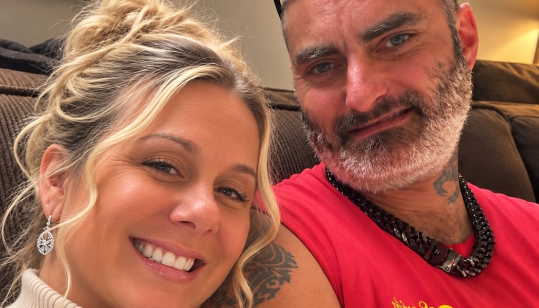 A smiling blonde woman and a bearded man in a red shirt sitting close together on a couch.