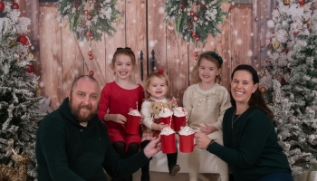 A smiling family of five holding red hot chocolate mugs in front of a snowy Christmas backdrop.