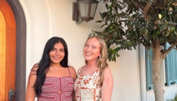 Two young women, one dark-haired and one blonde, smiling together outside a stucco building.