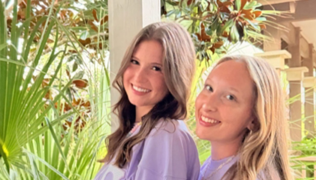 Two smiling young women in matching purple shirts pose outdoors in front of tropical greenery.