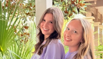 Two smiling young women in matching purple shirts pose outdoors in front of tropical greenery.