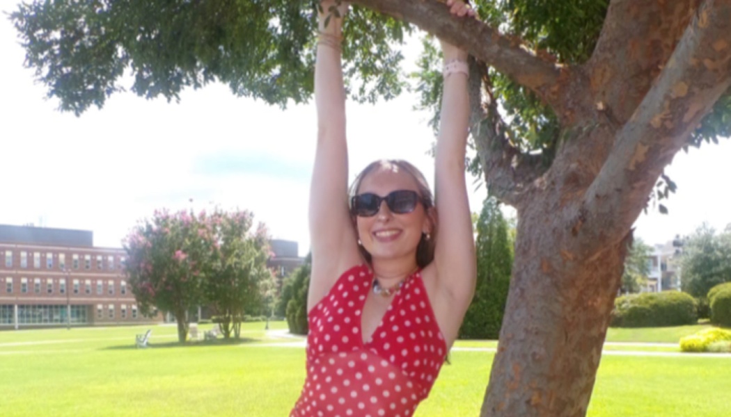 Smiling woman in sunglasses and a red polka dot dress hangs playfully from a tree branch on a sunny campus.