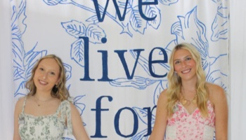 Two smiling young women in floral dresses pose in front of a white and blue banner reading "We live for".