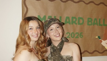 Two smiling young women in camo attire pose in front of a banner reading "Mallard Ball 2026."