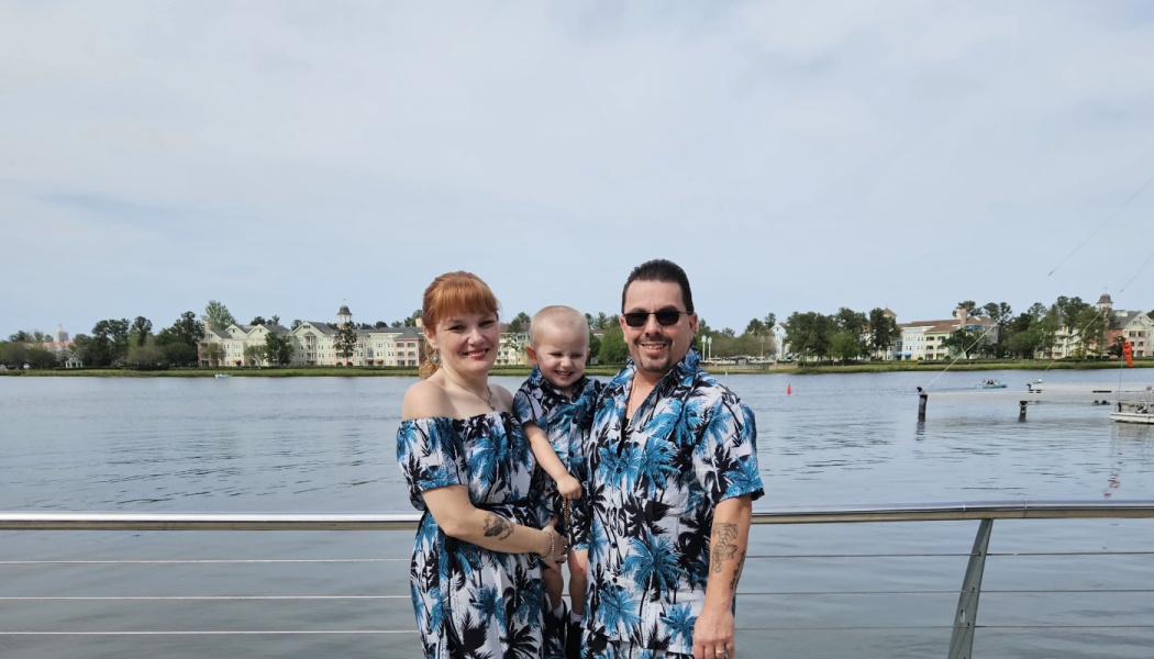 A smiling family of three wearing matching blue palm tree outfits outdoors in front of a scenic lake.