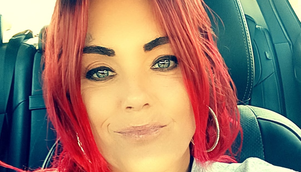 A woman with bright red hair, blue eyes, and hoop earrings smiling while sitting inside a car.