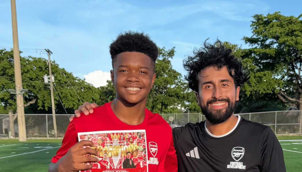 Two smiling young men in Arsenal soccer jerseys pose outdoors on a field, one holding a certificate.