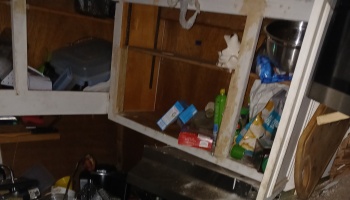Damaged, open wooden kitchen cabinets filled with scattered food boxes and household items.