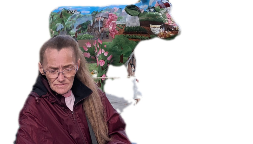 Woman with glasses in a maroon jacket looking down, in front of a colorful painted cow statue.