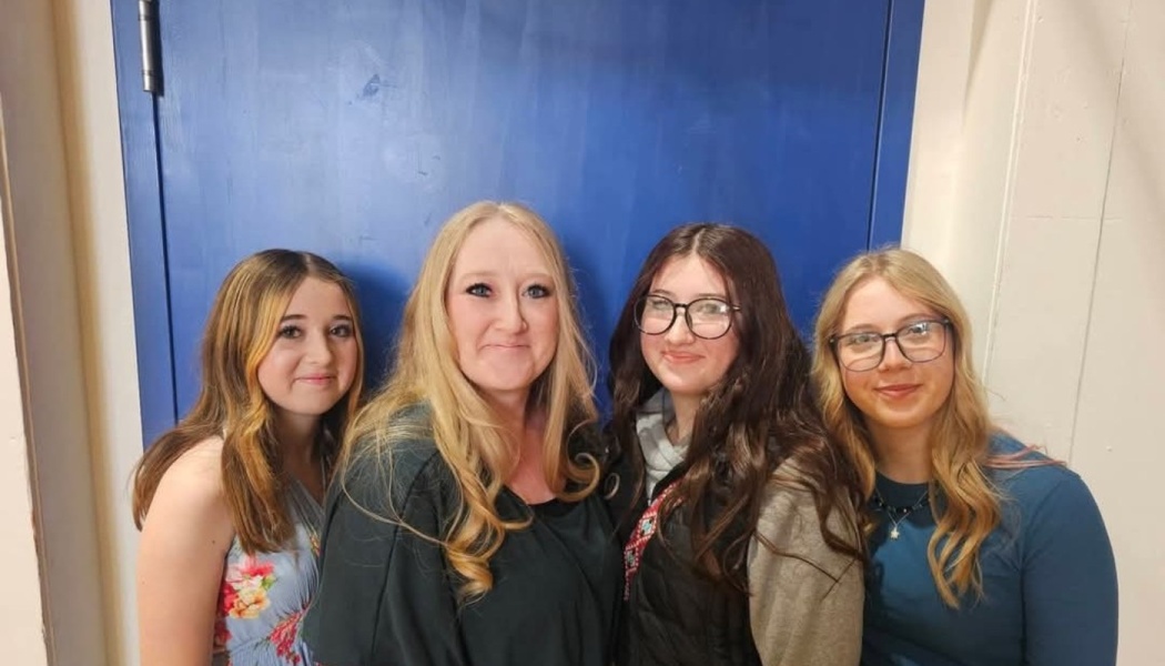 A blonde woman and three young women smiling together side-by-side in front of a blue door.