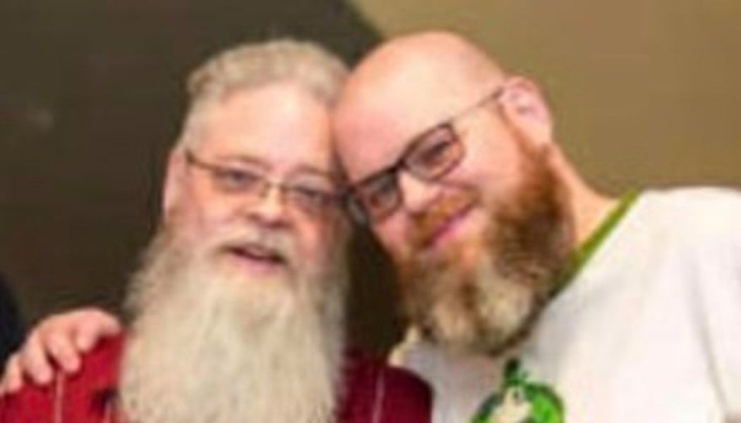 Two smiling men with glasses and full beards leaning their heads together affectionately.
