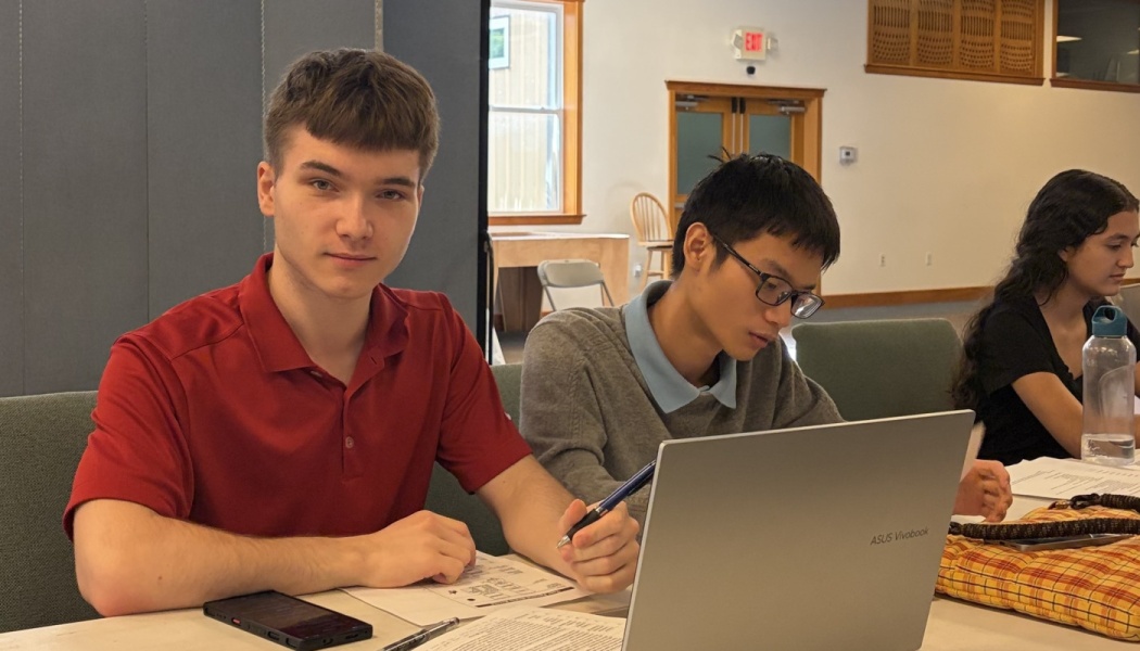A student in a red shirt looks at the camera while peers work on laptops and papers at a table.
