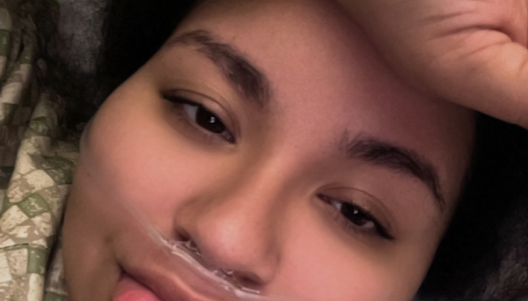 Close-up selfie of a young person lying in bed with a nasal cannula and a hand on their forehead.