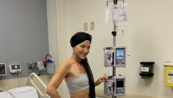 A smiling woman wearing a headscarf stands in a hospital room next to an IV drip pole.
