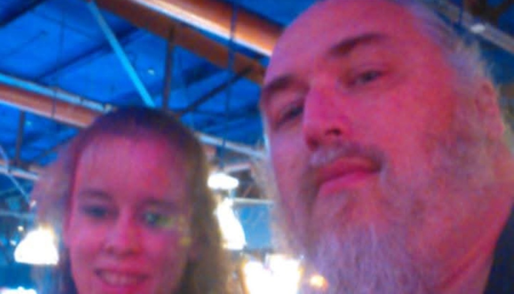 Close-up selfie of a bearded man with a smiling woman behind him, lit with vibrant pink and blue lights.