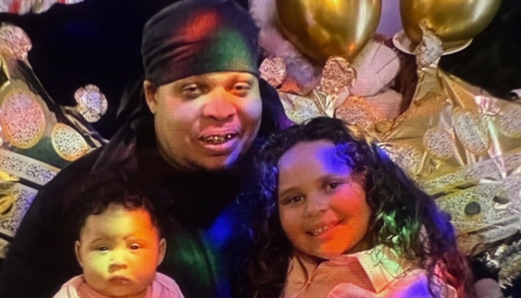 A smiling man poses with a baby and a young girl with curly hair in front of gold party balloons.