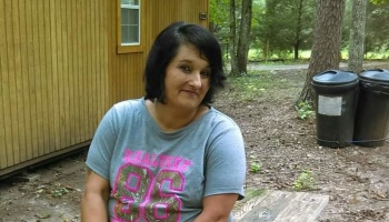A smiling woman with short dark hair wearing a gray graphic t-shirt sits outdoors near a wooden cabin.
