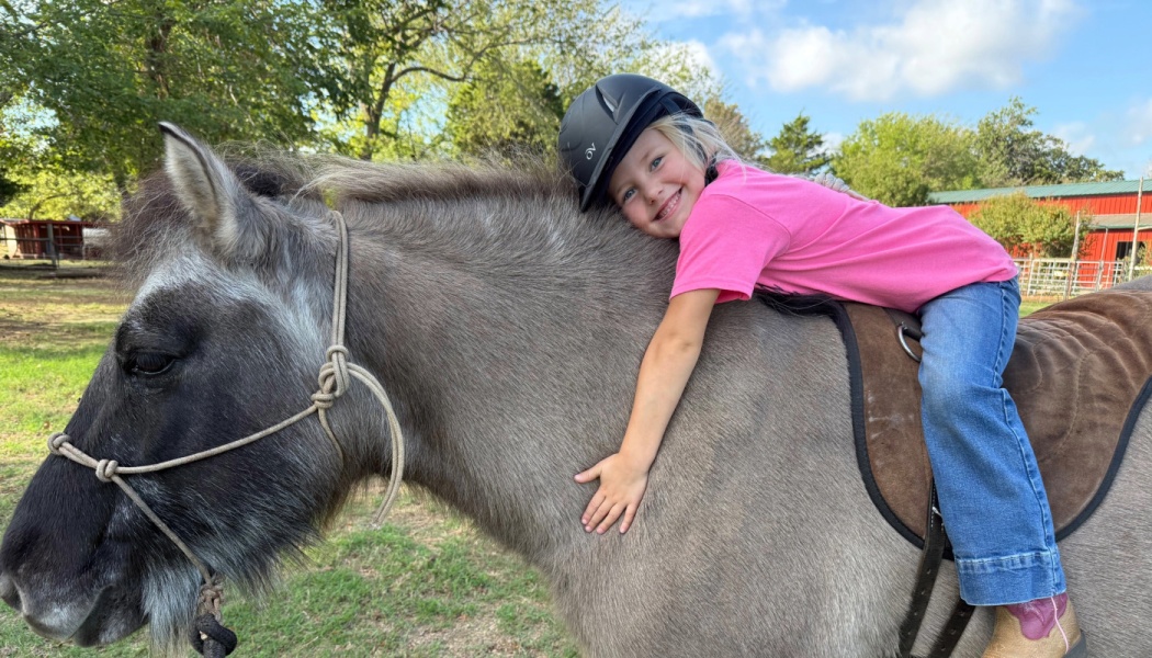 A smiling young girl in a pink shirt and riding helmet hugging a gray pony's neck outdoors.