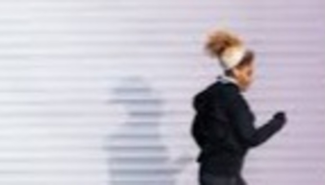 A woman in black athletic wear jogging past a white ribbed wall, casting a shadow behind her.