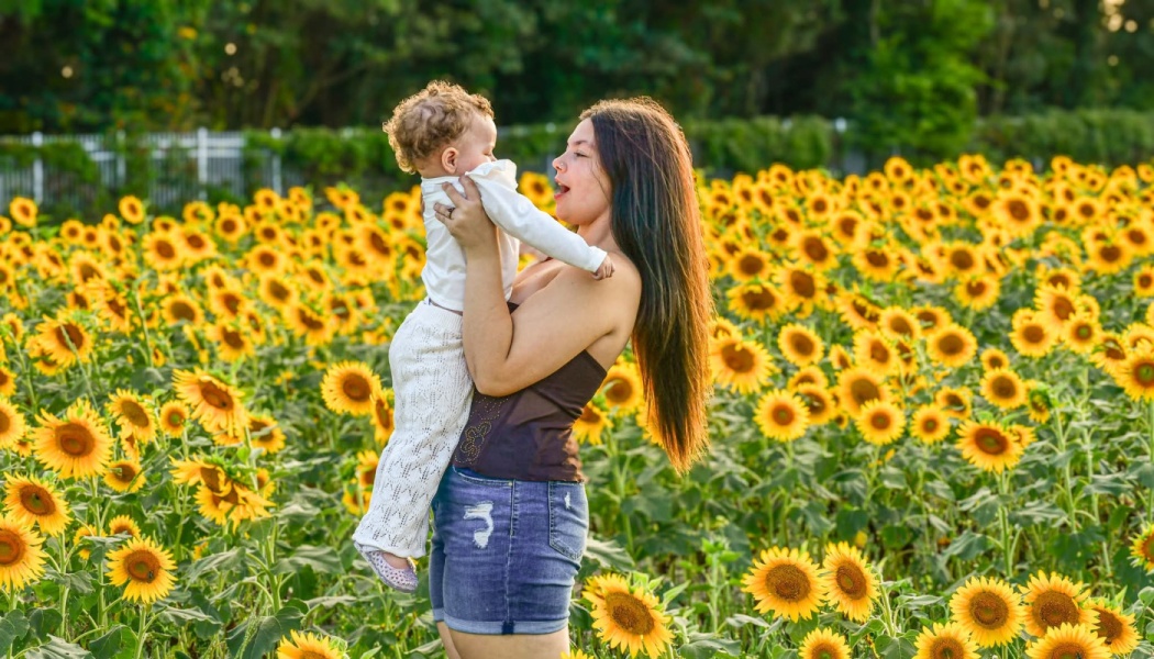 A mother holds her young child up in a bright field surrounded by blooming yellow sunflowers.