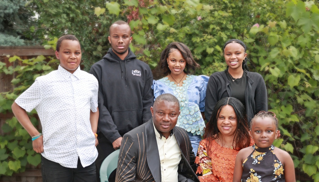 A smiling family of seven, including parents and five children, posing together outdoors in front of green foliage.