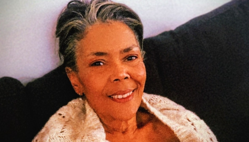 Smiling older woman with short gray hair wearing a cream knit sweater, sitting on a dark couch.