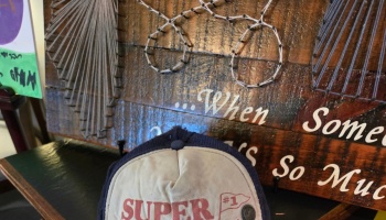 A baseball hat reading "SUPER #1" in front of a rustic wooden sign with string art and cursive text.