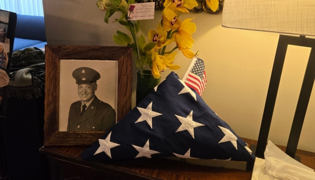 Framed military portrait, folded American flag, and yellow sympathy flowers displayed on a wooden table.