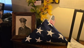 Framed military portrait, folded American flag, and yellow sympathy flowers displayed on a wooden table.