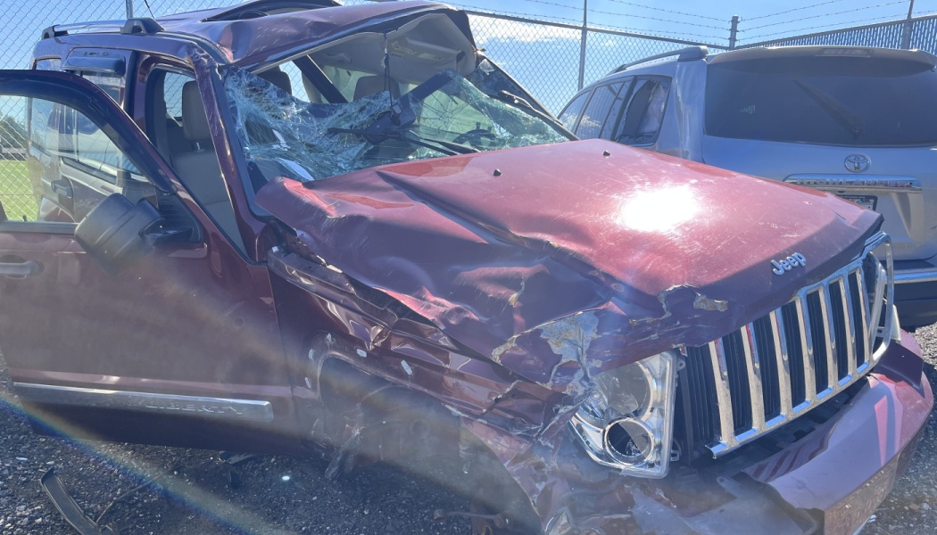 A heavily damaged maroon Jeep SUV with a crushed front end, smashed windshield, and caved-in roof.