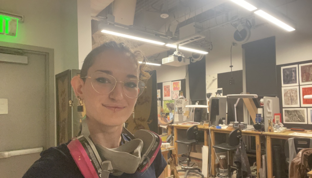 A smiling artist with glasses wearing a respirator mask around their neck in an art workshop.