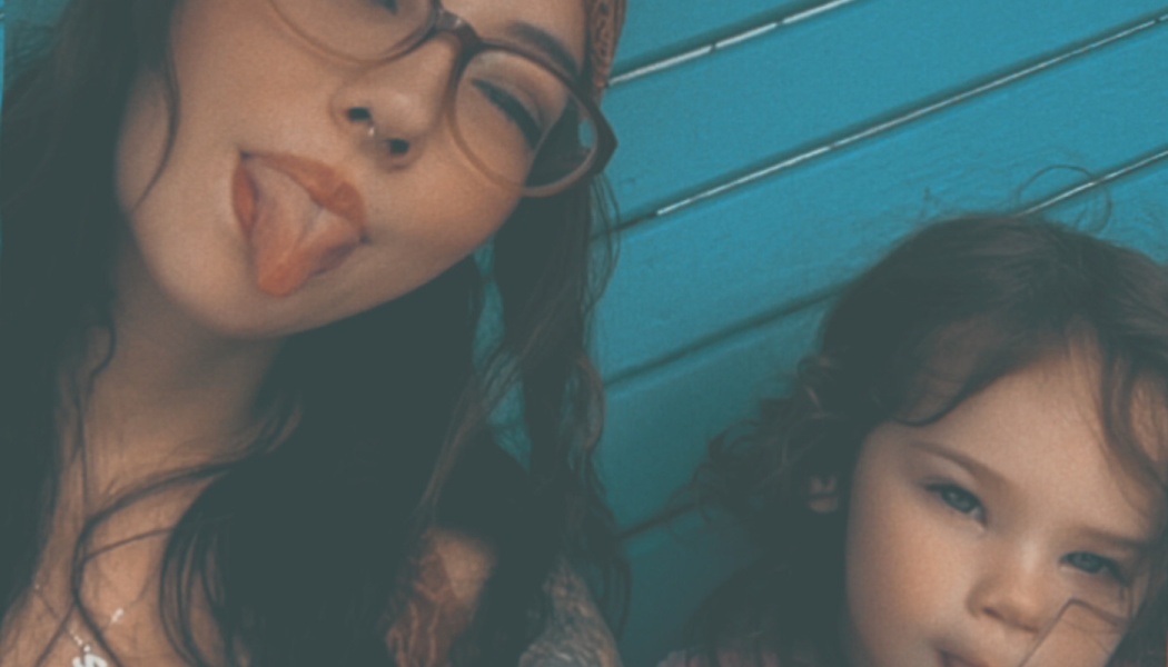 Playful selfie of a woman with glasses sticking out her tongue next to a young child in front of a blue wooden wall.