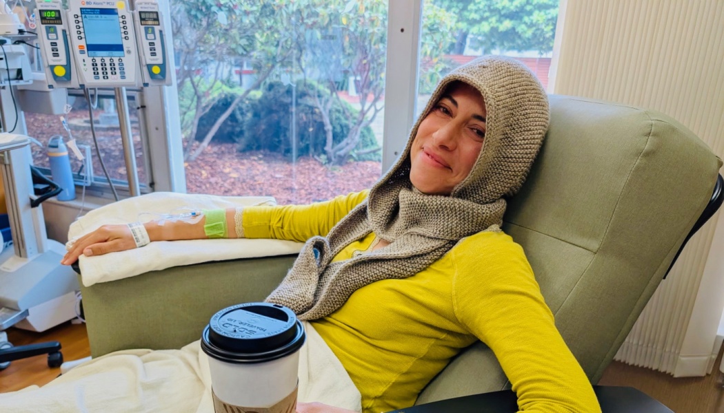 A smiling woman in a knit hood sits in a hospital recliner receiving IV treatment while holding a coffee cup.
