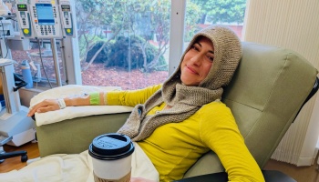 A smiling woman in a knit hood sits in a hospital recliner receiving IV treatment while holding a coffee cup.