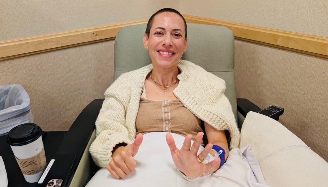 A smiling woman with a shaved head sits in a medical chair with an IV in her hand, giving a thumbs-up.