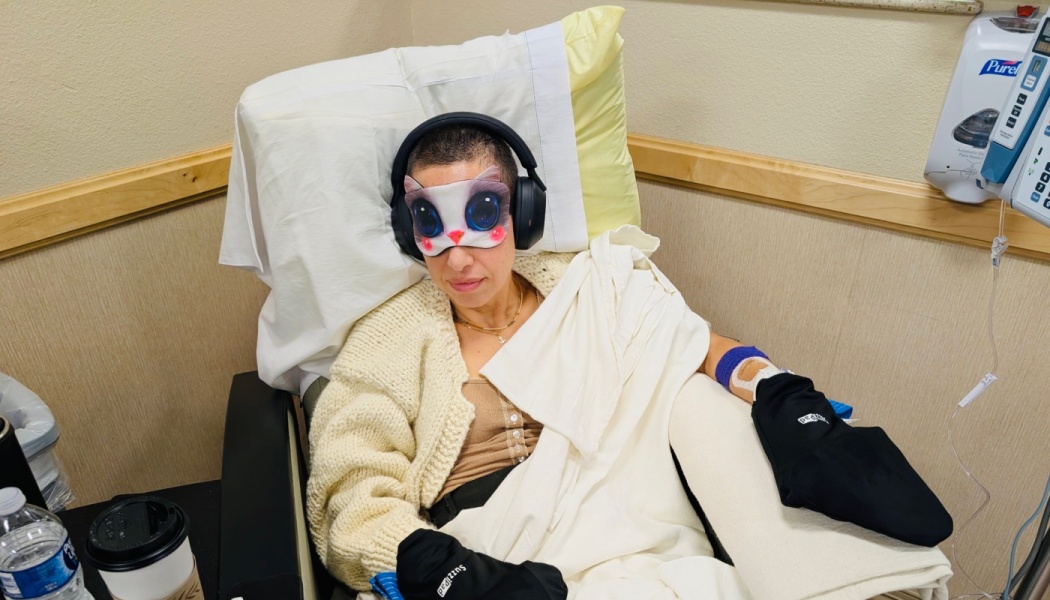 Woman with shaved hair in a medical chair wearing headphones, a cat eye mask, and cooling gloves.