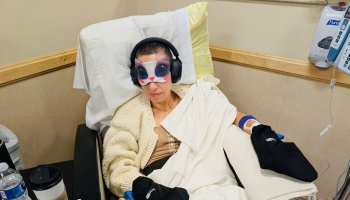 Woman with shaved hair in a medical chair wearing headphones, a cat eye mask, and cooling gloves.