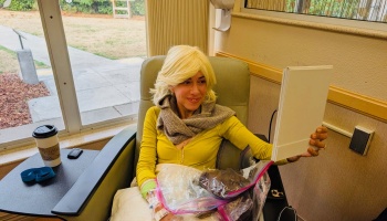 A smiling woman wearing a blonde wig looks at herself in a mirror while sitting in a medical treatment chair.
