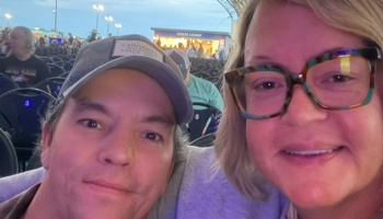 A man in a baseball cap and a blonde woman in glasses smile together in a close-up selfie at an outdoor venue.