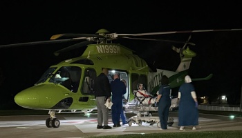 Medical personnel loading a patient on a stretcher into a green medical helicopter on a helipad at night.