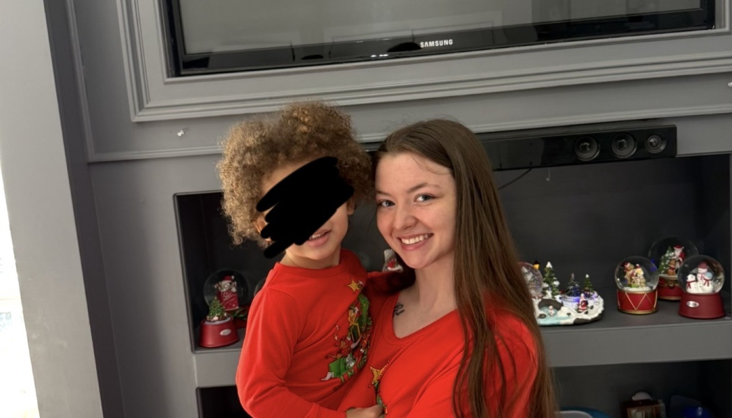 A smiling young woman holding a young child in matching red holiday tops, surrounded by festive Christmas decor.