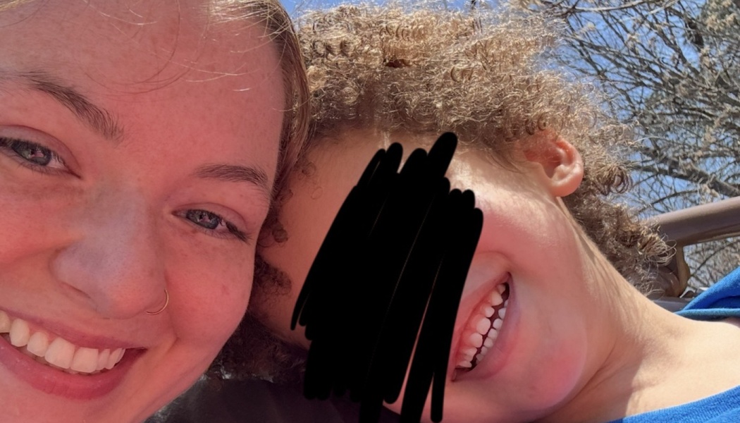 Close-up selfie of a smiling young woman and a happy curly-haired child posing together outdoors on a sunny day.