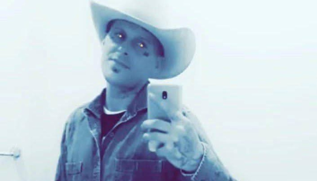 A man in a cowboy hat taking a mirror selfie with a smartphone in a blue-toned image.