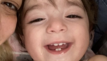 Close-up of a smiling toddler showing tiny front teeth, cuddled next to a parent.