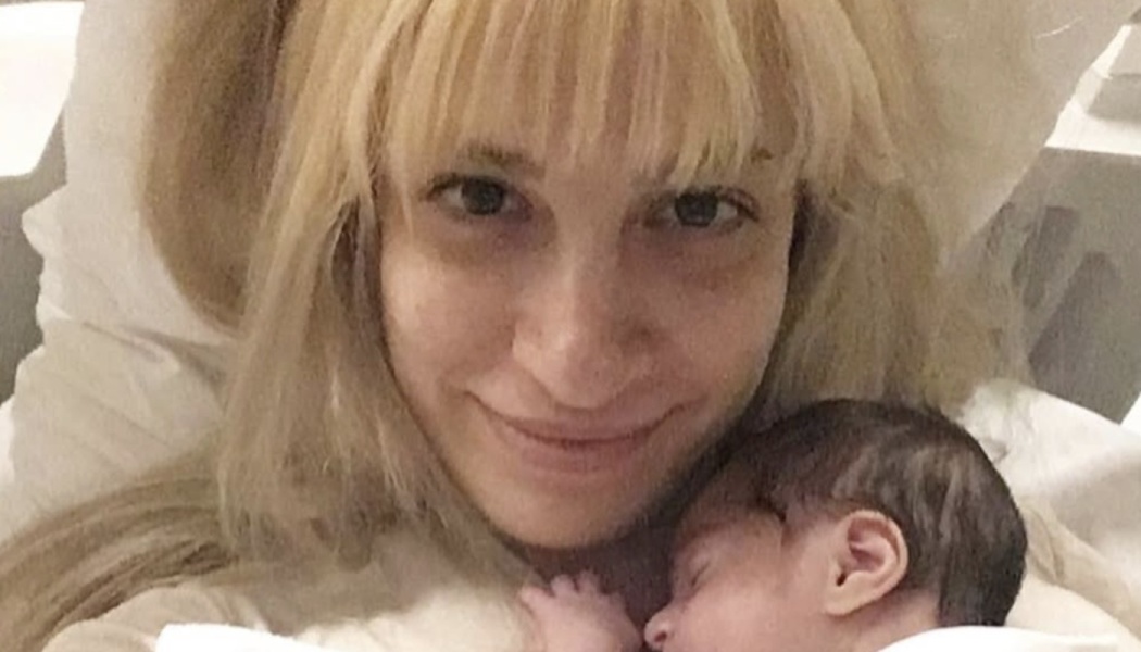 A smiling blonde mother lying in bed and holding her sleeping newborn baby close to her chest.