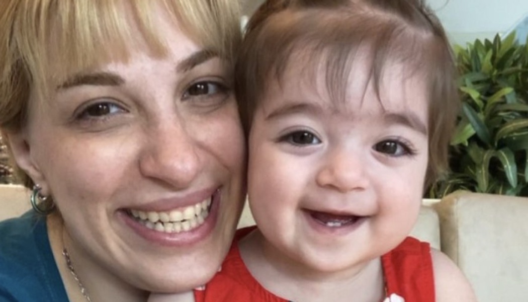 A smiling woman with blonde hair poses happily with a smiling baby girl wearing a red top.
