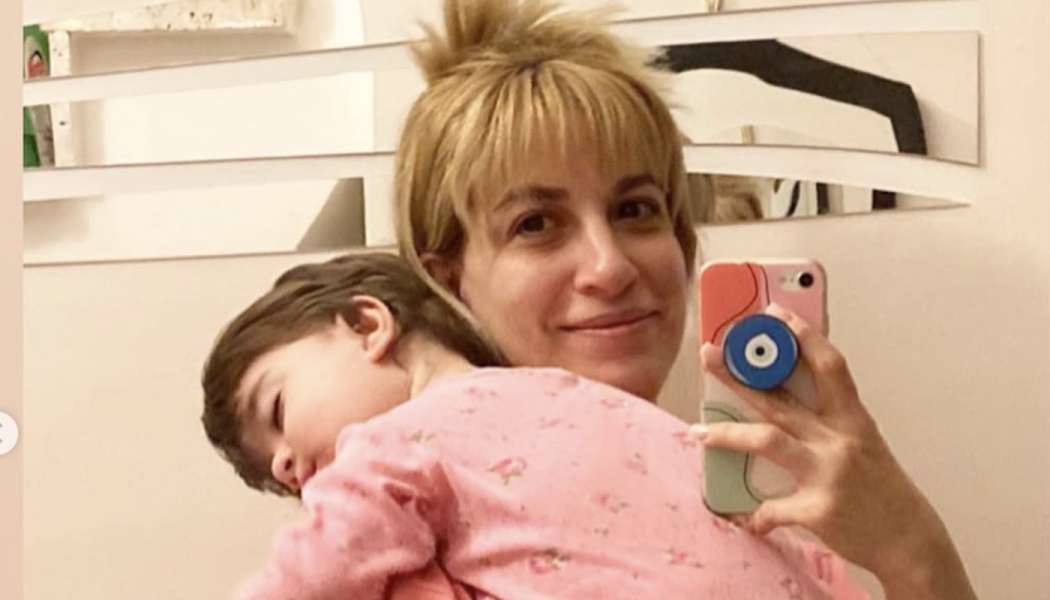 A blonde woman takes a mirror selfie while holding a sleeping toddler in pink pajamas on her shoulder.