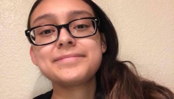 Young woman with dark hair and black rectangular glasses smiling gently against a plain wall.